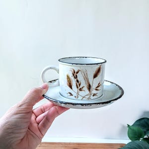 May include: A white ceramic coffee cup and saucer set. The cup and saucer have a brown rim and feature a wheat stalk design. The cup has a handle and is resting on the saucer.