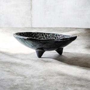 May include: A dark, oval-shaped ceramic bowl with three short legs. The bowl has a textured surface and a glossy, iridescent finish that reflects shades of blue, green, and purple. The bowl is a decorative piece.