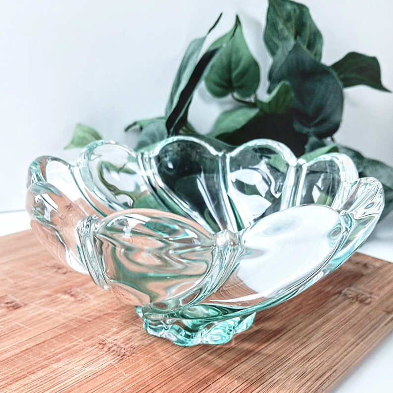 May include: A clear, scalloped-edge glass bowl with a light teal tint. The bowl sits on a wooden surface, with green foliage in the background. The bowl's design features a series of petal-like shapes, creating a decorative and elegant appearance.