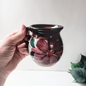 May include: A hand-painted ceramic mug with a dark brown rim and handle. The mug features a large, stylized burgundy flower with green leaves against a white background. The mug is held by a person's hand.
