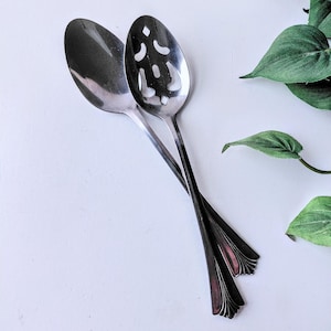 May include: Two stainless steel serving spoons. One is solid, the other has a slotted design. The spoons are angled, with handles overlapping. The handles have a decorative, fan-like design at the end. Green leaves are in the background.