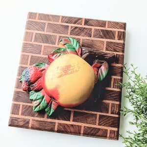 May include: A wooden cutting board with a brick pattern in brown and orange. A ceramic fruit decoration with a yellow peach and red strawberries is attached to the board.