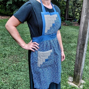 May include: A blue apron featuring a log cabin quilt pattern on the bib and pocket. The apron is constructed from a small-scale floral print fabric in shades of blue and cream, with solid blue fabric accents on the waistband and neck straps.