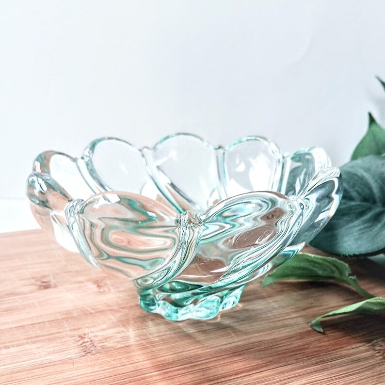 May include: A clear glass bowl with a scalloped edge, resembling a flower. The bowl has a pedestal base and sits on a wooden surface. The glass has a slight teal tint, and the bowl is empty.