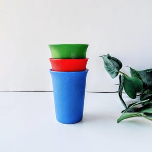 May include: Three colorful plastic cups stacked on top of each other. The bottom cup is blue, the middle cup is red, and the top cup is green. The cups are on a white surface with a green plant in the background.