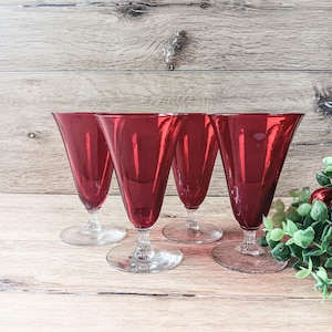 May include: Four red-colored stemmed glasses with a conical shape. The glasses have clear, circular bases and are arranged on a light-colored wooden surface. The background features a wooden wall.