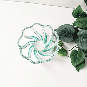 May include: A clear glass bowl with a spiral design featuring teal accents. The bowl has a scalloped edge and a white center. The bowl is on a white surface with green leaves in the background.