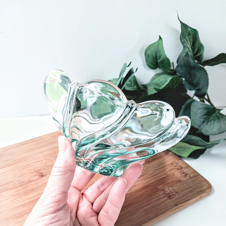 May include: A clear, decorative glass bowl with a unique, petal-like design. The bowl has a light green tint and is held over a wooden cutting board. The bowl's shape and color create a delicate and elegant aesthetic.