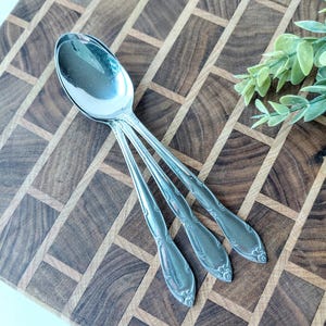 May include: Three silver spoons with ornate handle detailing are arranged on a wooden cutting board. The spoons have a classic design with a rounded bowl and decorative patterns. The wooden board has a geometric pattern.
