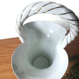 May include: White ceramic vase with a gold-trimmed handle. The handle is twisted and has a decorative gold stripe design. The vase has a wide, open mouth and a narrow base.