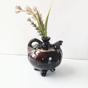 May include: A brown ceramic pitcher with a spout and three legs. The pitcher has a floral design and is filled with pink and green flowers.