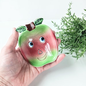 May include: A ceramic apple-shaped container with a smiling face. The apple is green and red with blue eyes, a brown stem, and green leaves. The container is held in a hand.
