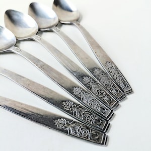 May include: A set of five silver-toned metal spoons arranged in a fan pattern. Each spoon features a long, flat handle decorated with an embossed floral motif consisting of a rose and leafy foliage at the base of the handle stem.