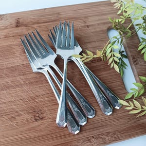 May include: A set of eight silver forks with ornate designs on the handles. The forks are stacked on a wooden cutting board.
