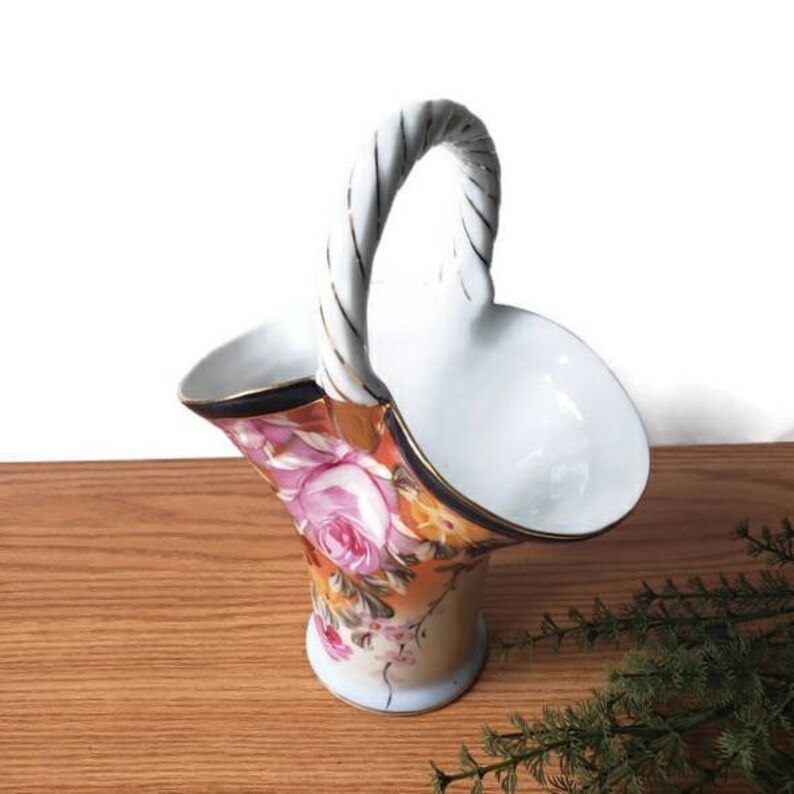 May include: A white porcelain basket with a twisted rope handle and floral design. The basket is decorated with pink, yellow, and orange flowers on a white background.