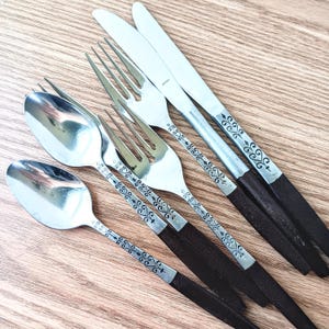 May include: A set of five silver-toned flatware pieces with black handles. The pieces include two spoons, two forks, and one knife. The handles have a decorative floral pattern.