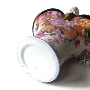 May include: A vintage porcelain vase with a floral pattern in shades of pink, orange, and green. The vase has a white base with a blue rim and a gold-coloured handle.