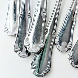 May include: Close-up of several silver-colored spoons with ornate, curved handles. The spoons are arranged at various angles, reflecting light and creating highlights. The design features a decorative border along the edges of the spoon handles.