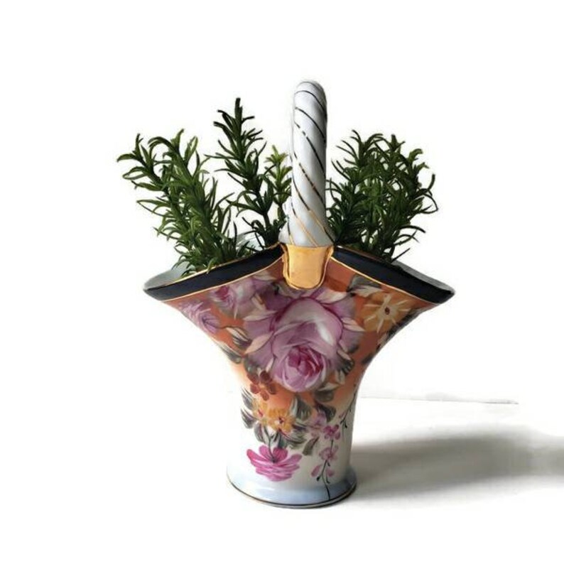 May include: A white porcelain vase with a floral pattern and a twisted handle. The vase is shaped like a basket and has a gold trim. The vase is filled with green leafy plants.