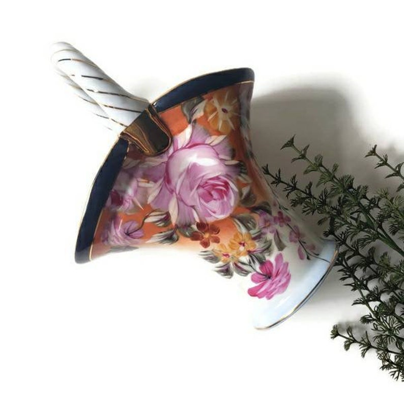 May include: A white porcelain vase with a twisted white handle. The vase is decorated with a floral pattern in shades of pink, orange, yellow, and blue.