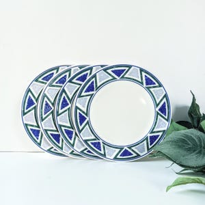 May include: Four white ceramic plates with a geometric design. The plates have a blue, green, and gray triangle pattern around the rim. The plates are stacked and slightly overlapping, with a white background.