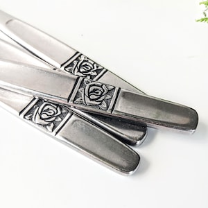 May include: Close-up of several silver-colored utensils. Each utensil has a rectangular design with a rose in the center. The utensils are arranged on a white surface, with some overlapping. The utensils are made of metal.