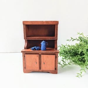 May include: A miniature wooden hutch with two doors and a shelf. The hutch is painted brown and has a blue ceramic teapot and two cups on the shelf.