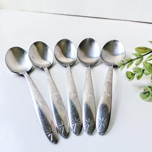 May include: A set of five silver-toned metal spoons arranged in a row. Each spoon features a smooth, rounded bowl and a handle decorated with a repeating engraved starburst or sunburst pattern along the length of the stem.