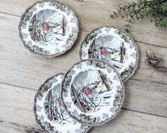 May include: Four white ceramic plates with a winter scene design. Each plate features a detailed illustration of a snowy landscape with trees, a red building, and figures. The plates have a scalloped edge with a decorative border.