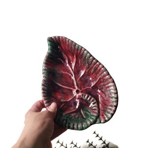 May include: A ceramic leaf-shaped dish with a deep red and green glaze. The dish has a raised, textured design that resembles a leaf.