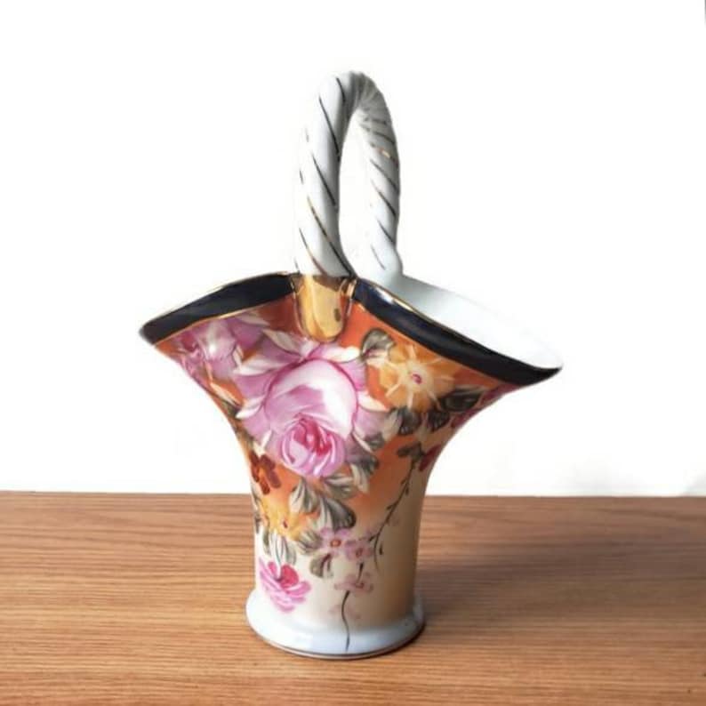 May include: A small white porcelain basket with a twisted rope handle. The basket is decorated with a floral pattern in shades of pink, yellow, and orange.