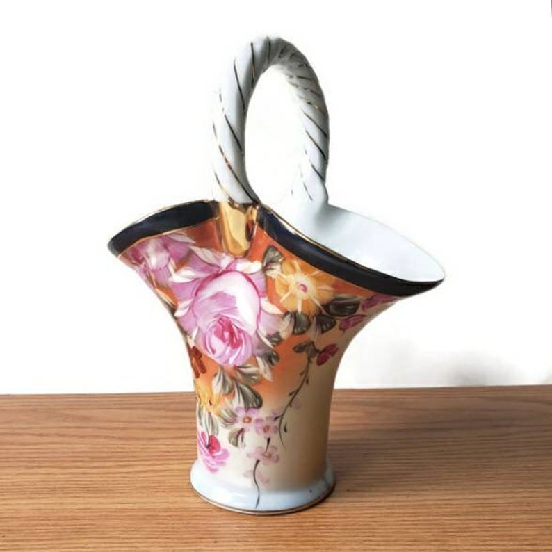 May include: A white porcelain vase with a twisted rope handle. The vase is decorated with a floral pattern of pink roses, yellow flowers, and green leaves on an orange background. The rim of the vase is trimmed with a gold and blue band.