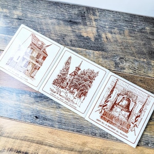 May include: Three white square coasters with brown illustrations of historical Philadelphia landmarks. The coasters feature images of a house, a church, and the Liberty Bell. Text on the coasters includes the names of the landmarks.