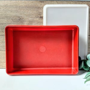 May include: A red rectangular plastic container with a matching lid. The container has a smooth surface and a small circular indentation in the center of the base. A white rectangular lid is behind the red container.