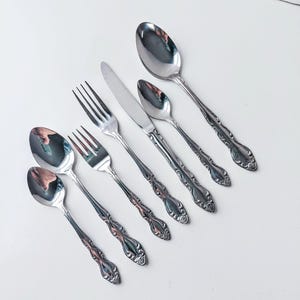 May include: A set of stainless steel silverware, including spoons, forks, and a knife, arranged on a white surface. The silverware has ornate, decorative handles. The spoons are of different sizes, and the forks have four tines.