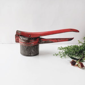 May include: A red and rusty metal potato masher with a handle and a round, perforated bowl.