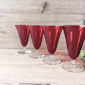 May include: Four ruby red wine glasses with a conical shape and clear stems and bases. The glasses are arranged in a row on a light brown wooden surface. The glasses are empty and reflect the light.