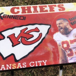 Op de afbeelding: Een rode, witte en zwarte Kansas City Chiefs tafel met een foto van Travis Kelce, nummer 87, aan de zijkant. De tafel heeft de woorden "Kennedi" en "Kansas City" erop.