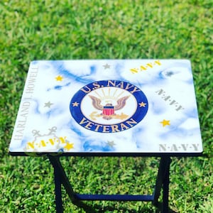 Op de afbeelding: Een witte opklaptafel met een blauw en gouden U.S. Navy Veteran-ontwerp. De tafel heeft een wit marmerachtig oppervlak met gouden sterren en de woorden "U.S. Navy Veteran" in blauwe en gouden letters. De tafel is ingeklapt en heeft een zwart metalen frame.