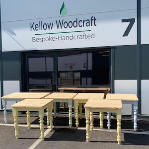 May include: A group of six wooden tables with turned legs in a light blue and yellow paint finish. The tables are arranged in a row in front of a building with a sign that reads "Kellow Woodcraft Bespoke-Handcrafted".