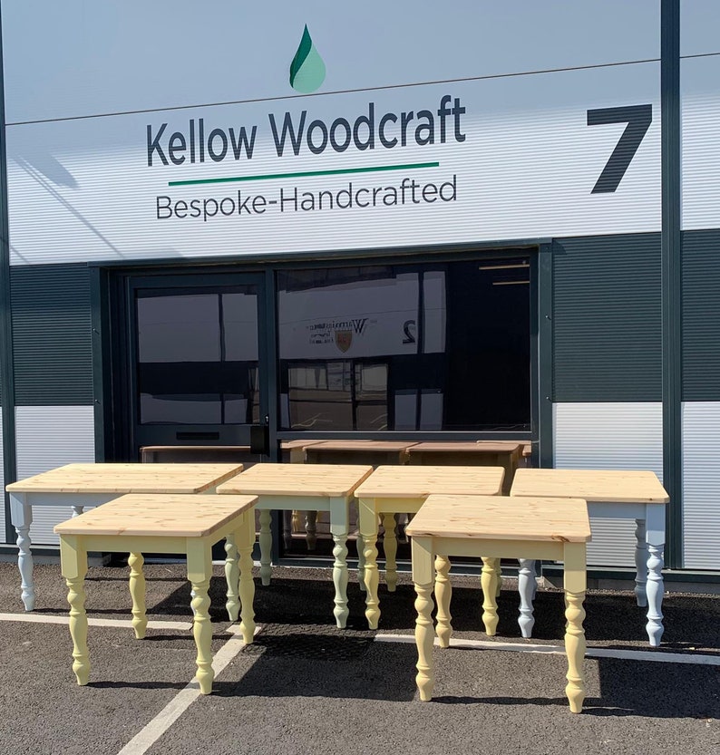 May include: A group of six wooden tables with turned legs in a light blue and yellow paint finish. The tables are arranged in a row in front of a building with a sign that reads "Kellow Woodcraft Bespoke-Handcrafted".
