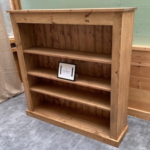May include: A rustic wooden bookcase with four shelves. The bookcase is made of natural wood with a light brown finish. The shelves are evenly spaced and have a slight lip to prevent items from falling off. The bookcase has a simple design with a flat top and a base with a slight overhang. The bookcase is standing on a gray carpet.