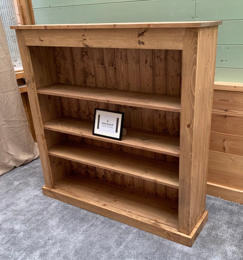 May include: A rustic wooden bookcase with four shelves. The bookcase is made of natural wood with a light brown finish. The shelves are evenly spaced and have a slight lip to prevent items from falling off. The bookcase has a simple design with a flat top and a base with a slight overhang. The bookcase is standing on a gray carpet.