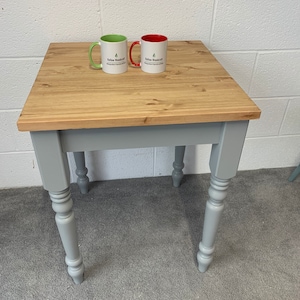 May include: A square wooden table with a natural light brown top and four grey painted legs featuring turned detailing. Two ceramic mugs sit on the surface, one with a green handle and one with a red interior, both printed with Kellow Woodcraft Bespoke-Handcrafted.
