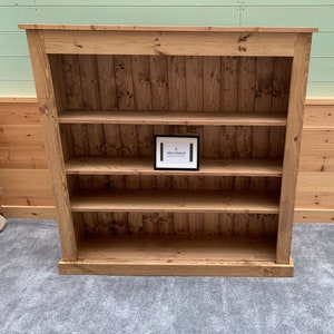 May include: A wooden bookcase with four shelves. The bookcase is made of natural wood with a light brown finish. The shelves are evenly spaced and have a slight lip to prevent items from falling off. The bookcase is standing on a gray carpet.