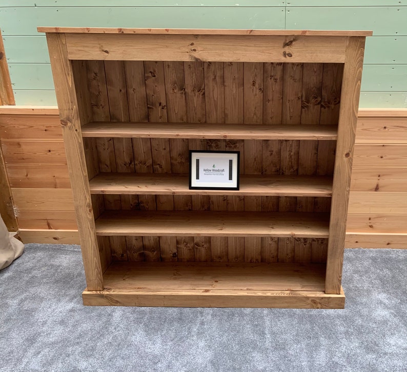 May include: A wooden bookcase with four shelves. The bookcase is made of natural wood with a light brown finish. The shelves are evenly spaced and have a slight lip to prevent items from falling off. The bookcase is standing on a gray carpet.