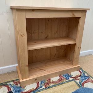May include: A natural wood bookshelf with two shelves. The bookshelf has a simple design with a base and a top trim.