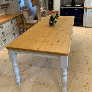 May include: A large, rectangular wooden table with a natural wood finish and white painted legs. The table has a turned leg design and is shown in a kitchen setting.