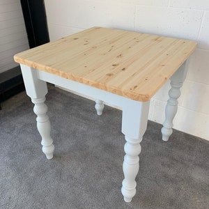 May include: A square wooden table with a light wood top and white painted legs. The legs have a turned design.