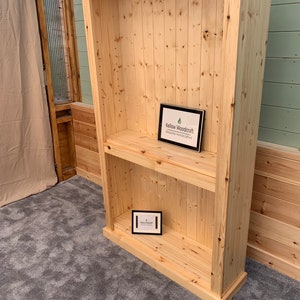 May include: A tall, wooden bookcase with two shelves. The bookcase is made of natural wood and has a simple, rustic design. The bookcase is empty, but there is a small sign on the bottom shelf that says "Kellow Woodcraft - Bespoke Handcrafted".